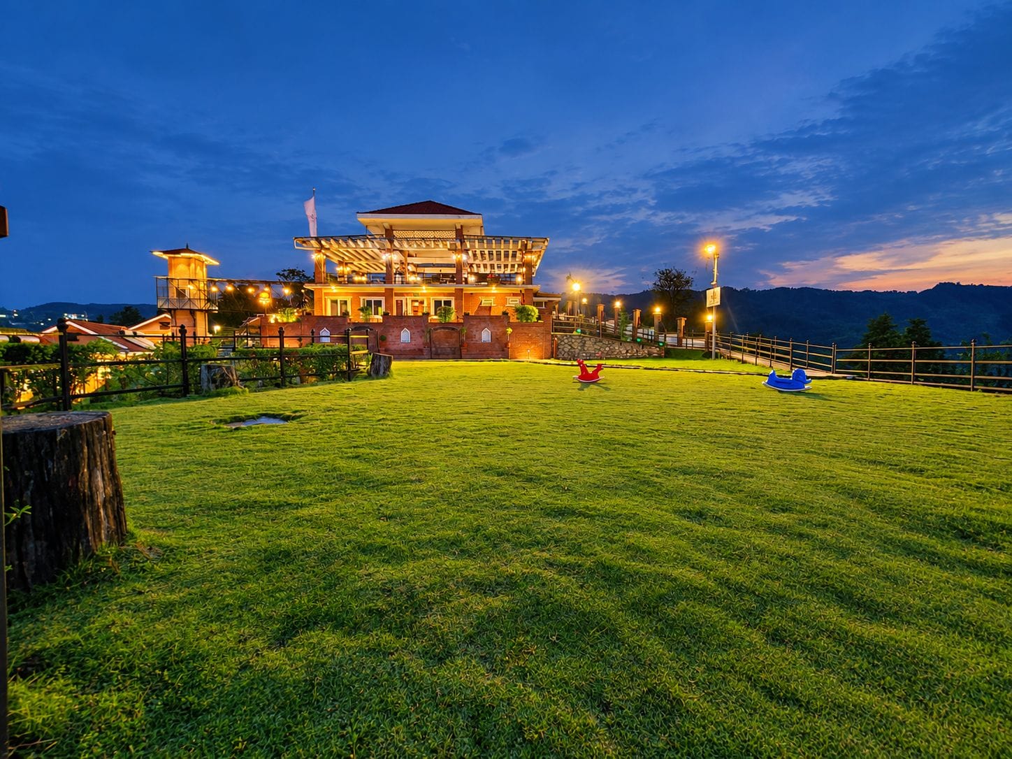 Un hotel bien iluminado con un amplio césped, una zona de juegos infantiles y montañas al fondo.