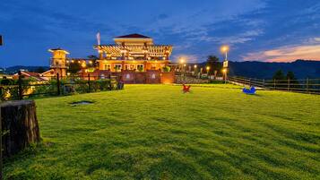 Un hotel bien iluminado con un amplio césped, una zona de juegos infantiles y montañas al fondo.