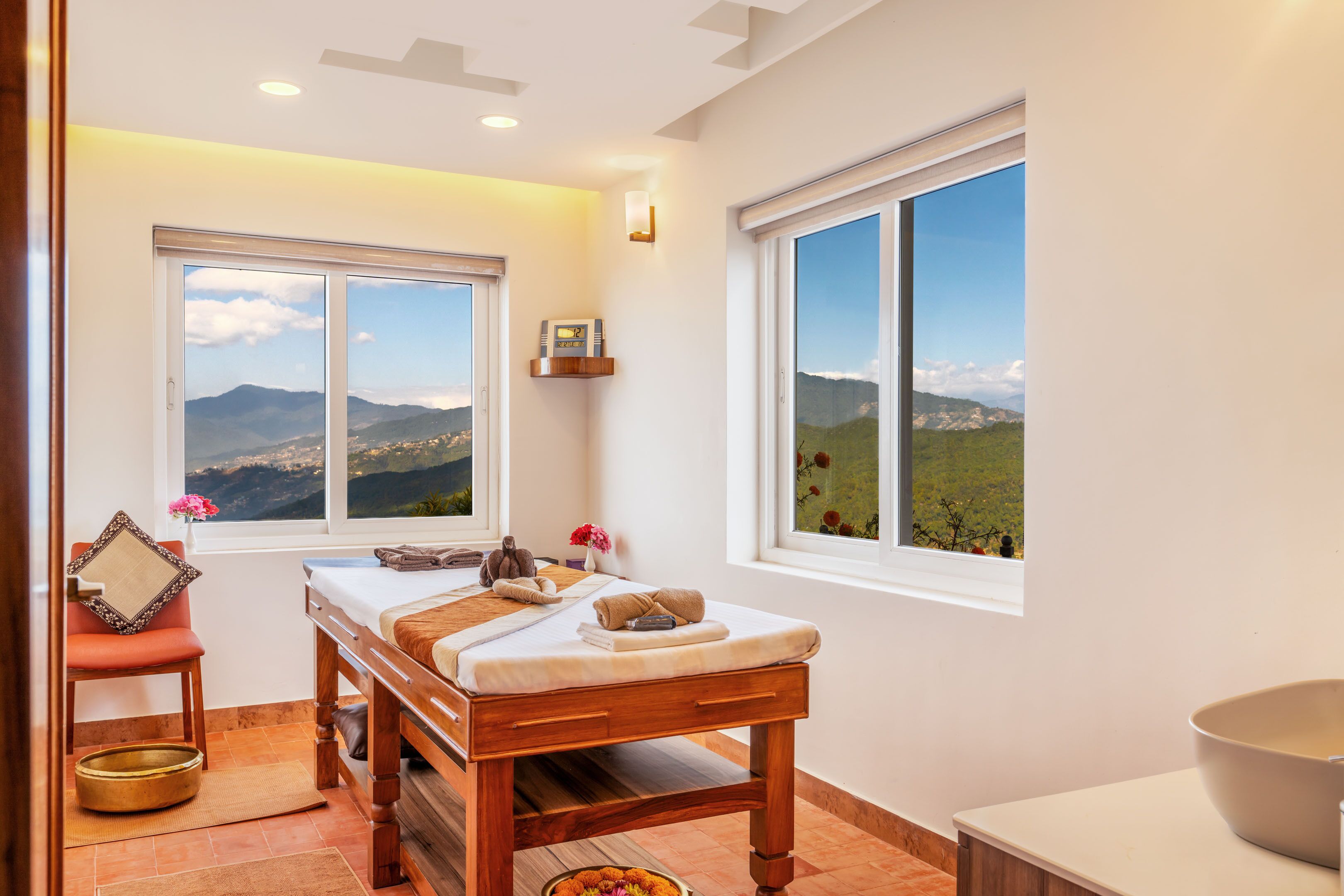 A spa room with a wooden massage table, two large windows with mountain views, a small shelf with a book, and a chair with a cushion.