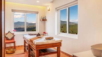A spa room with a wooden massage table, two large windows with mountain views, a small shelf with a book, and a chair with a cushion.