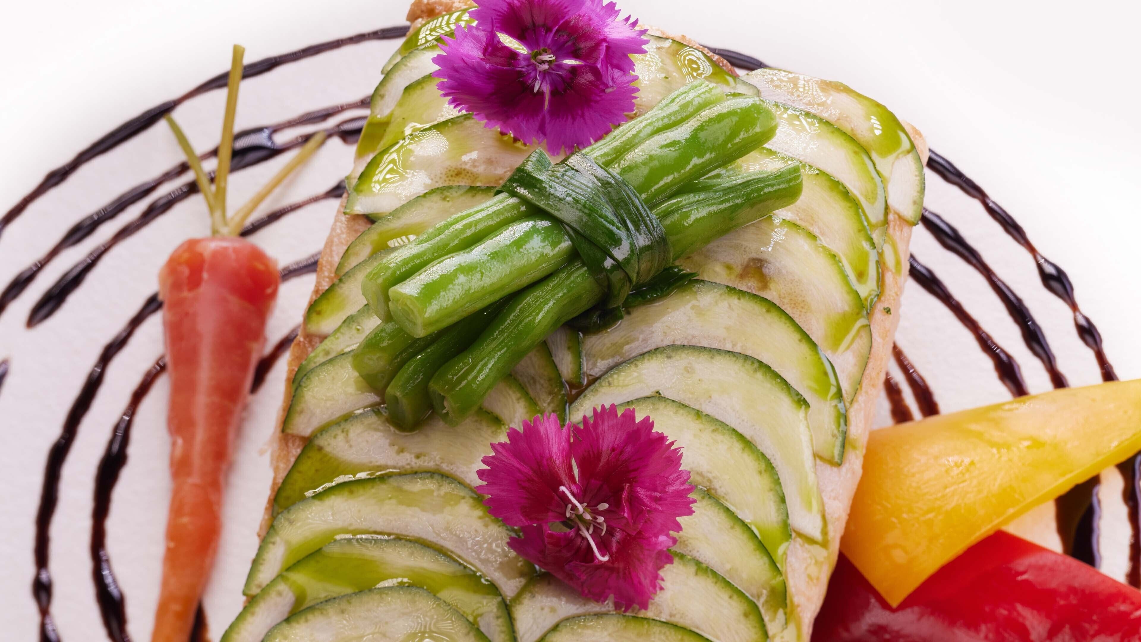 Un plat composé de rondelles de concombre, d’asperges vertes et de fleurs violettes, présenté sur une assiette blanche ornée de lignes noires.