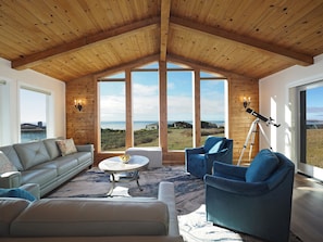 A spacious living room with a high wooden ceiling, large windows offering a view of the landscape, a telescope, and comfortable seating.