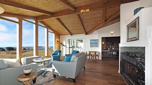 A spacious living area with a fireplace, wooden ceiling beams, and large windows offering a view of the outdoors. - Spacious * Ocean View * Walking Trail to Beach * Hot Tub (Bodega Bay)