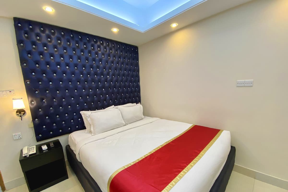 A hotel room with a tufted headboard, a bed with white bedding and a red and gold accent, a bedside table with a phone and a lamp, and a ceiling light.