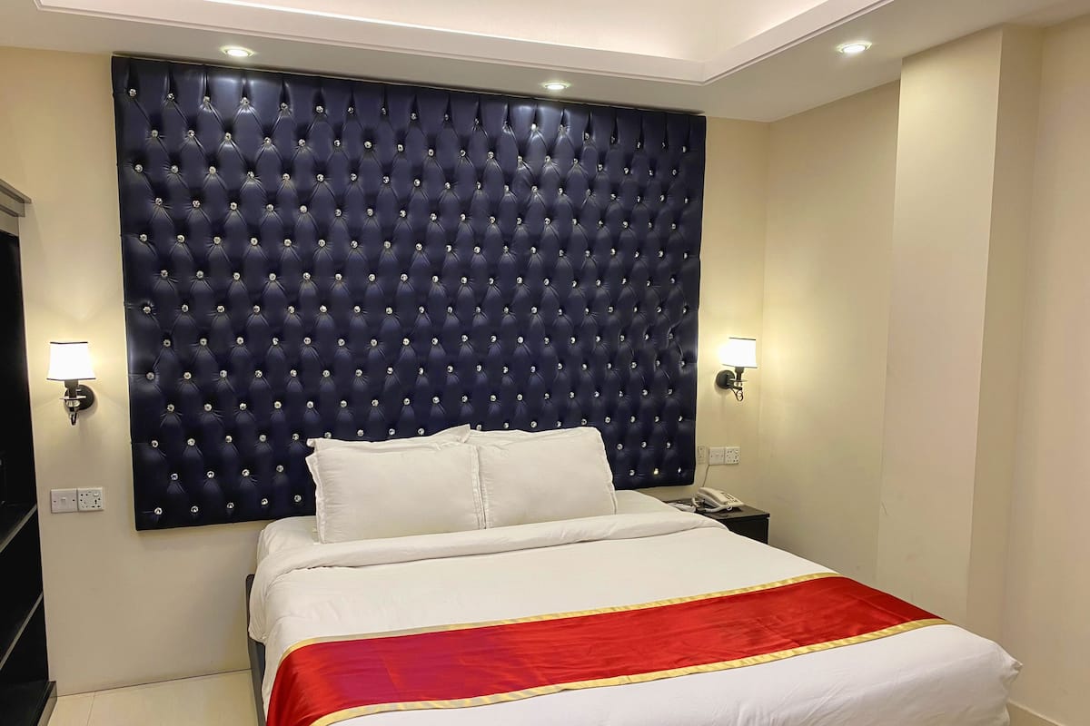 A hotel room with a tufted headboard, a bed with white linens and a red and gold accent, two bedside lamps, and a telephone on the nightstand.