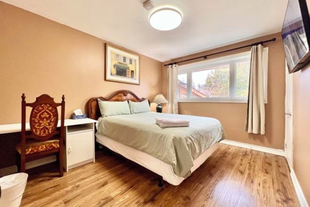 Un dormitorio con cama, mesita de noche, silla, ventana con cortinas e imagen en la pared.
