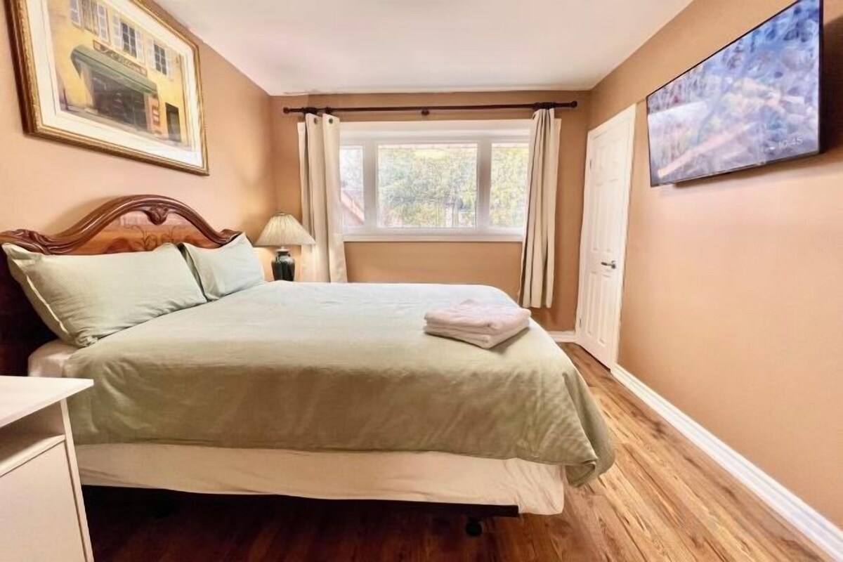 Un dormitorio con cama, mesita de noche, lámpara, ventana con cortinas y un cuadro en la pared.