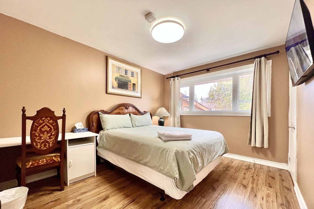 A bedroom with a bed, a chair, a nightstand, a lamp, a window with curtains, and a painting on the wall.