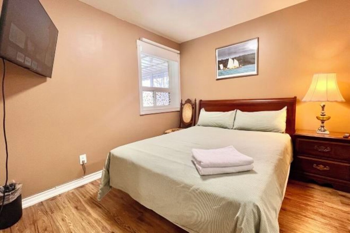 Un dormitorio con cama, mesita de noche, lámpara, un cuadro y ventana.