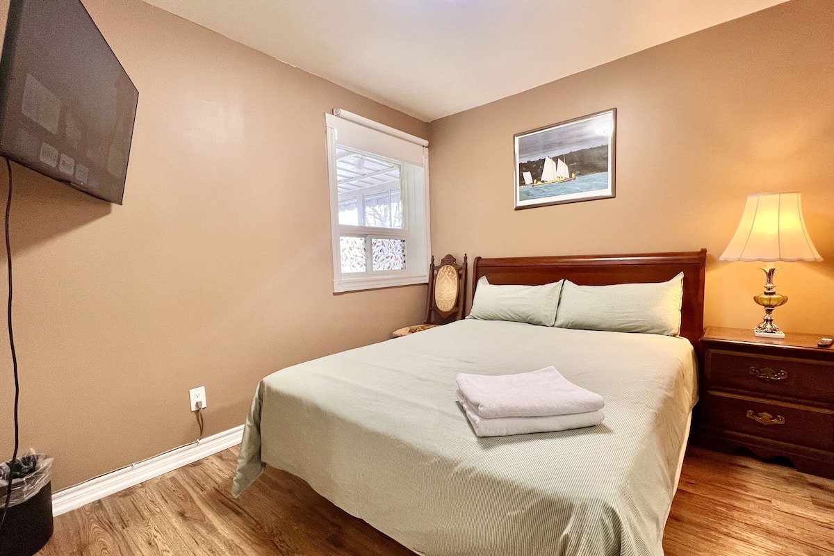 A bedroom with a large bed, a nightstand with a lamp, a window with blinds, and a painting on the wall.