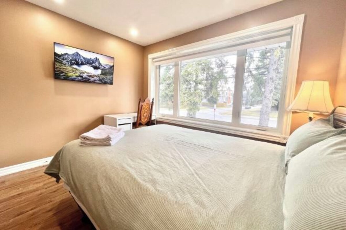 Un dormitorio con una cama grande, una ventana con vistas a árboles, un cuadro en la pared y una mesita de noche con una lámpara.