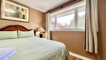 Una cama bien hecha con cabecera, lámpara de noche, ventana con cortinas e imagen enmarcada en la pared.