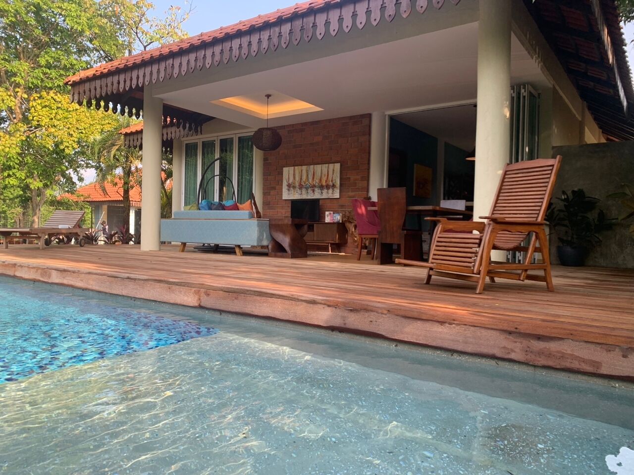 A swimming pool with a wooden deck, a covered seating area, and a brick wall with a painting.
