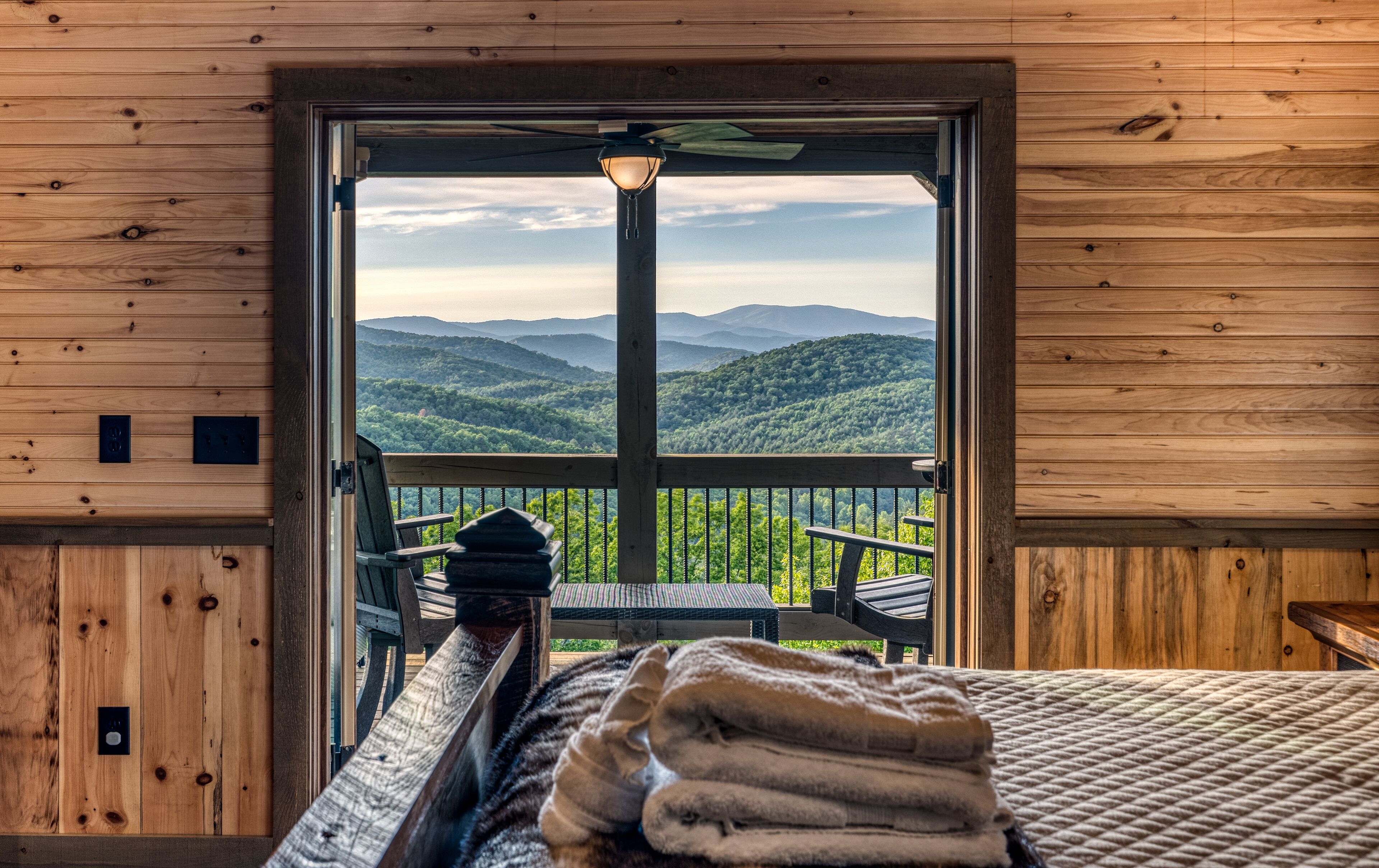 A wooden cabin room with a bed, a chair, and a view of mountains through an open door.