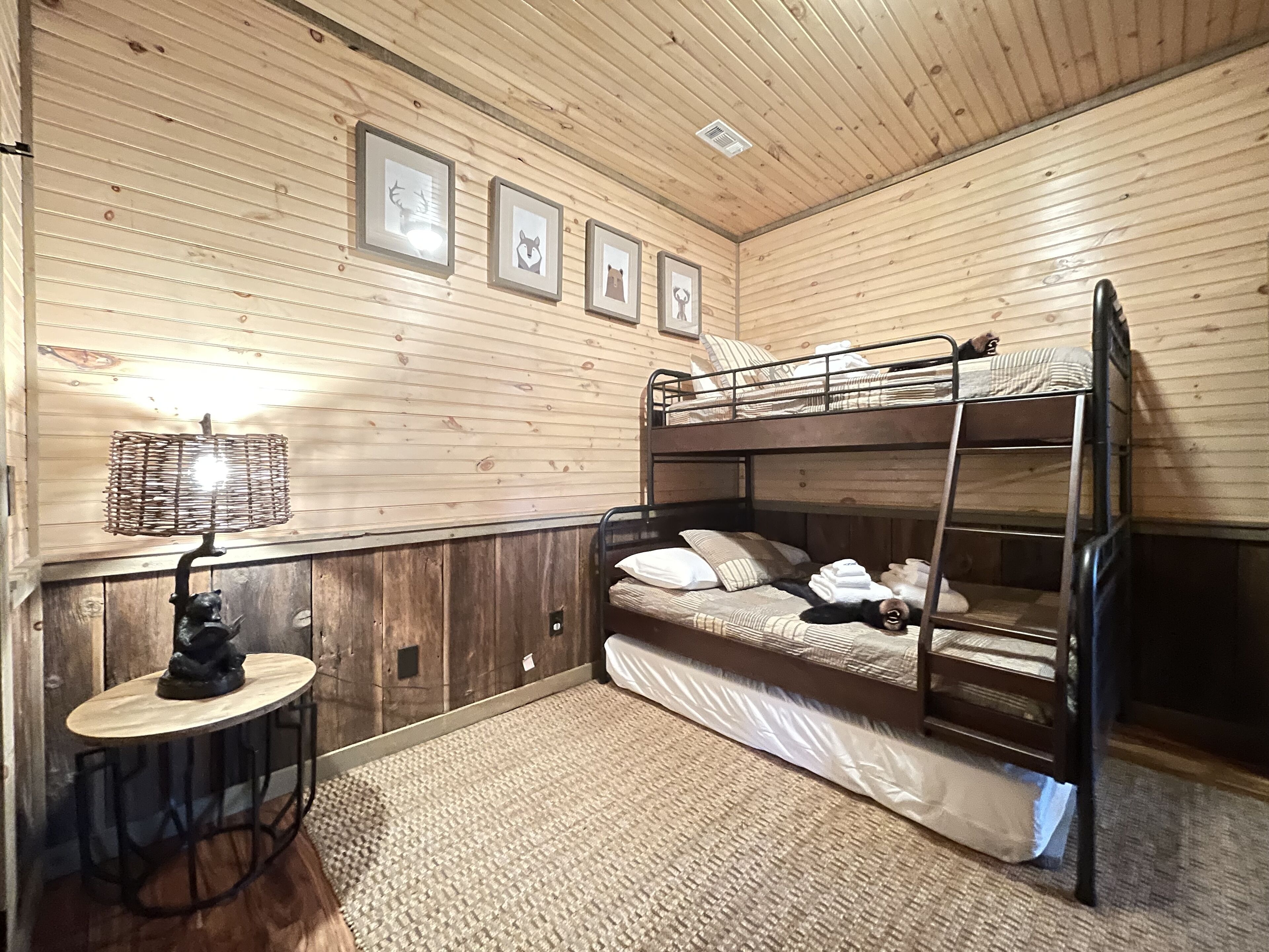 A bunk bed room with wooden walls, a small table with a lamp, and framed pictures on the wall.