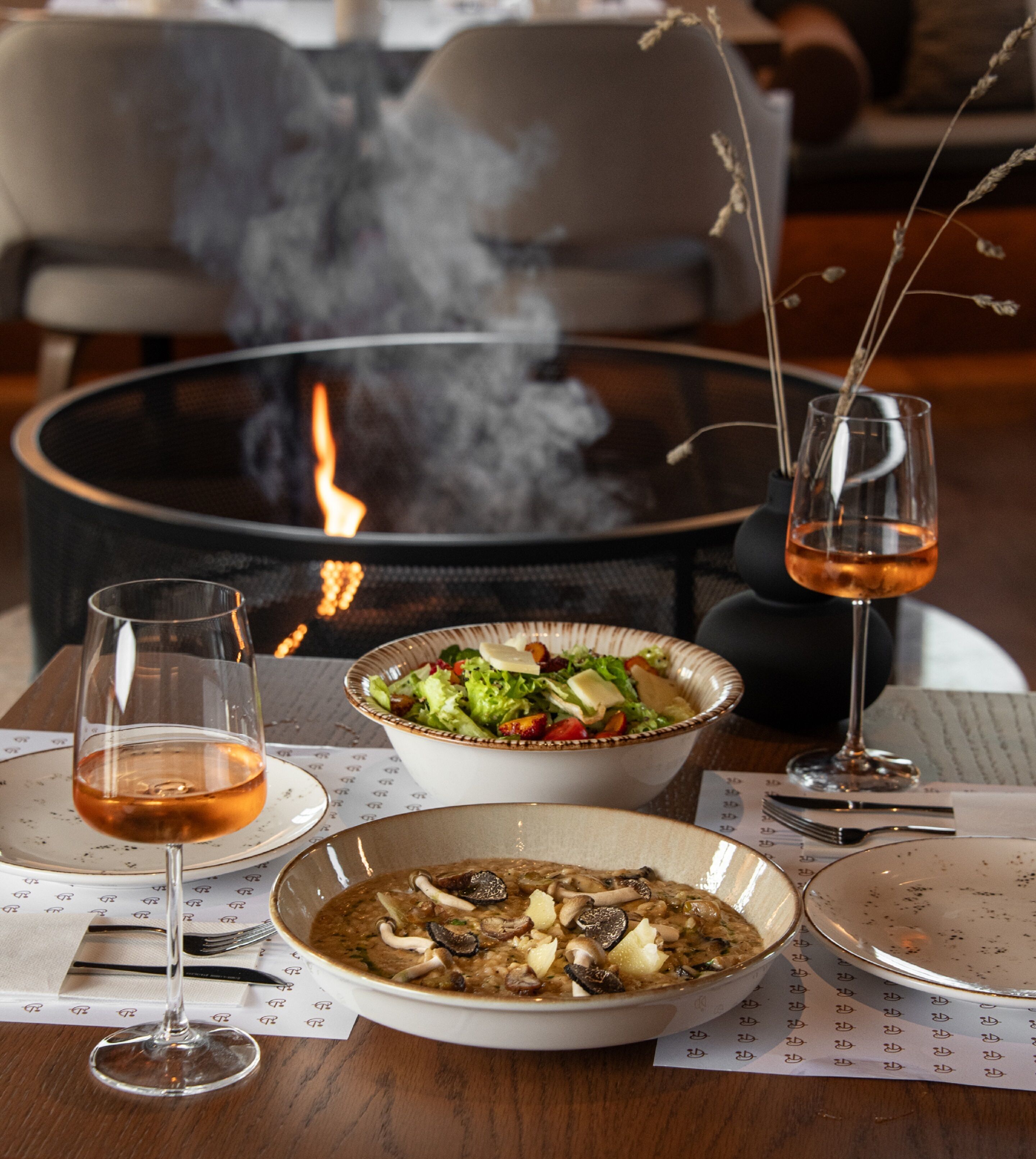 Uma mesa de jantar com lareira, dois copos de vinho e uma saladeira.