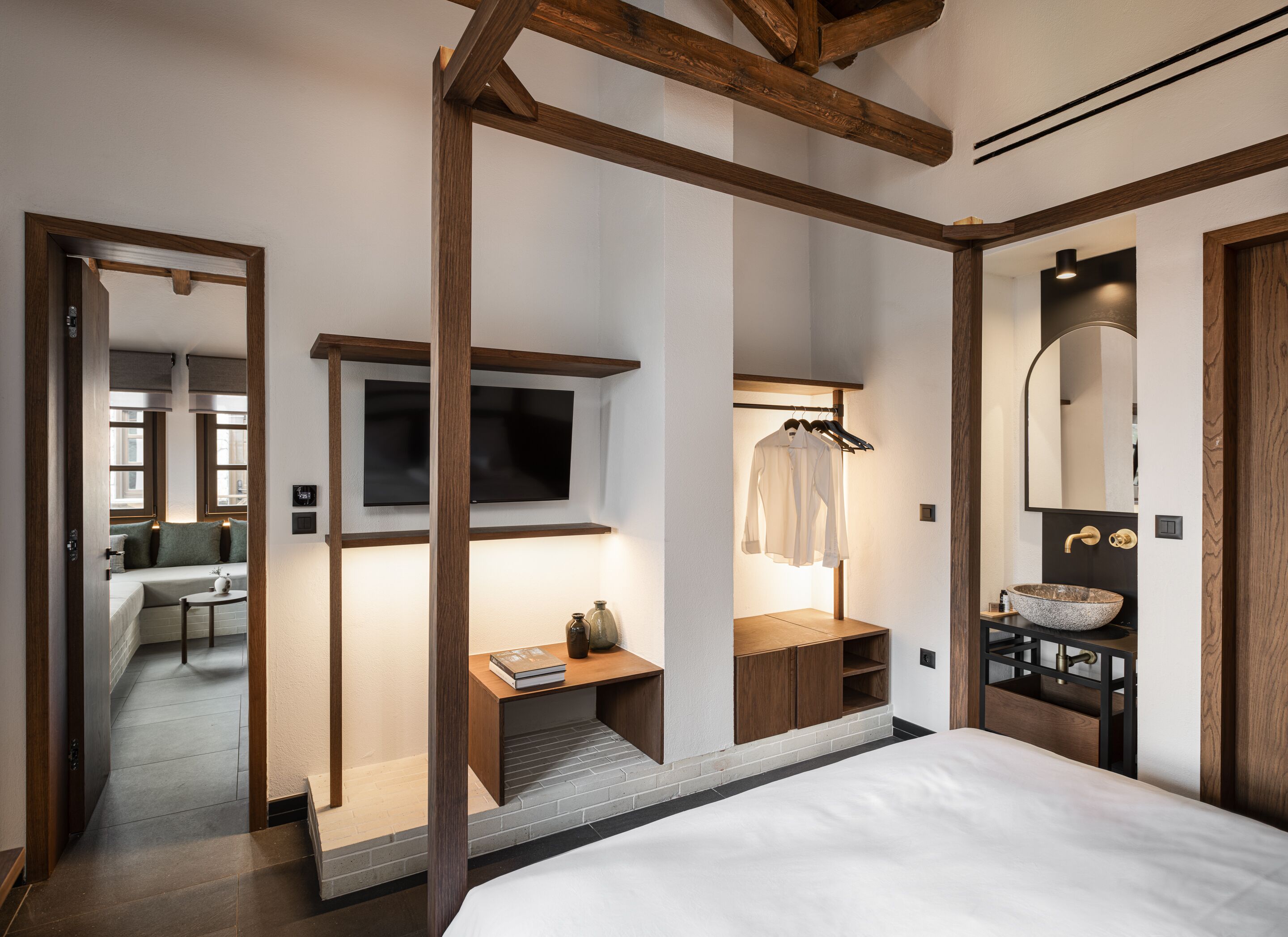 Ein modernes Schlafzimmer mit einem großen Bett, einem an der Wand befestigten Fernseher, einem Holztisch und einem Badezimmer mit einem großen Spiegel und Waschbecken.