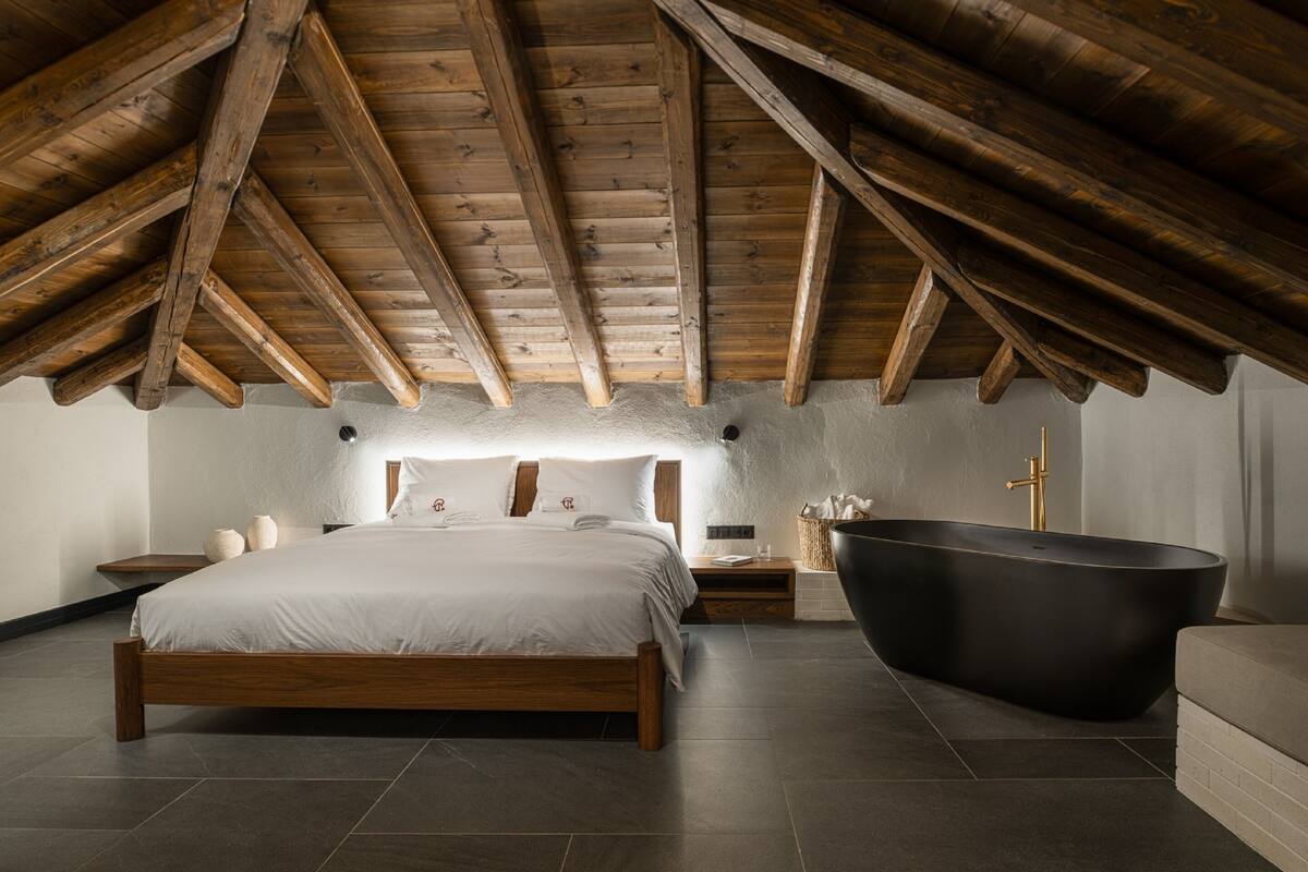 Un dormitorio con techo de madera, una cama grande, una bañera negra y una mesita de noche con lámpara.