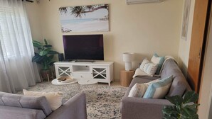 A living room with a grey sofa, a smaller grey armchair, a white entertainment unit with a flat-screen TV, a round side table, and a large potted plant. - Sunset Lakeview. (Windang)