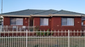 A single-story brick house with a dark tiled roof, a white picket fence, and a small porch with a bench.