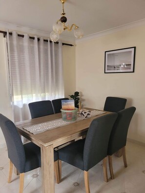 Un coin repas avec une table en bois, six chaises, un lustre, un tableau encadré au mur et une fenêtre avec des rideaux.