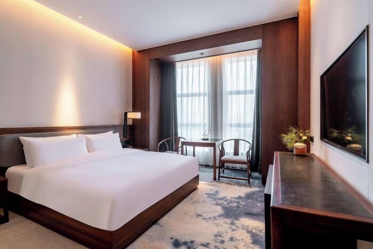 A hotel room with a large bed, a desk, a chair, a TV, and a window with curtains.
