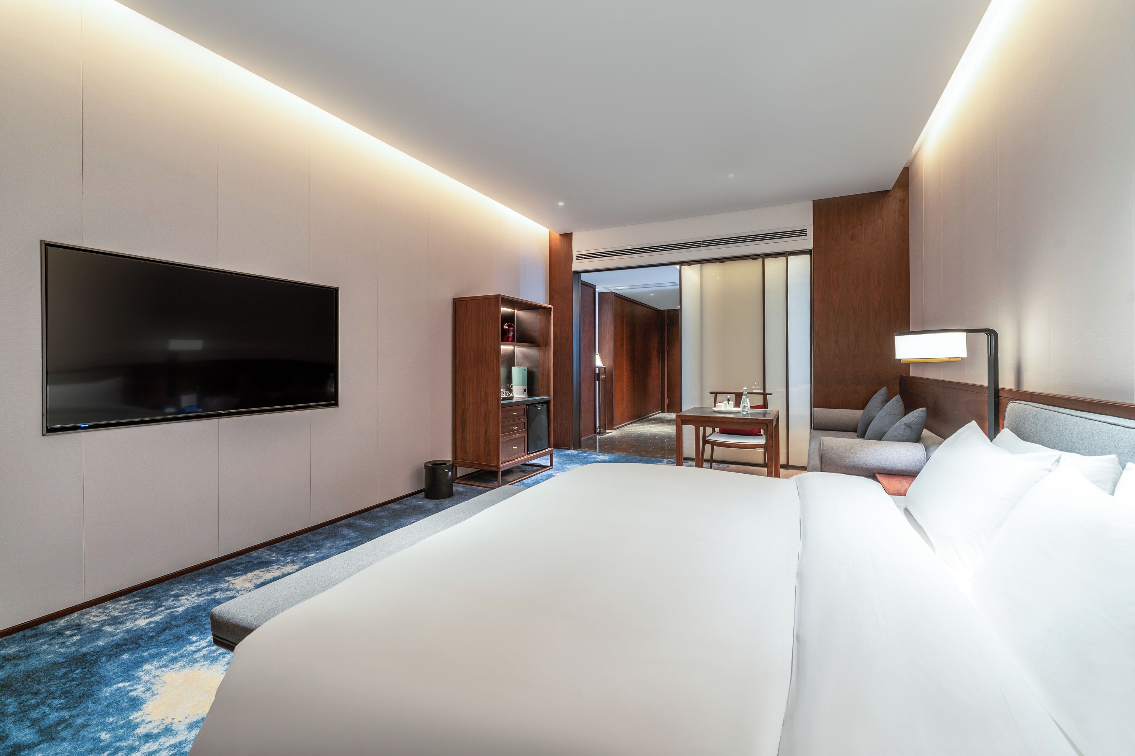 Una habitación de hotel con una cama grande, un televisor de pantalla plana montado en la pared, un armario de madera, una mesita con una silla y un espejo.