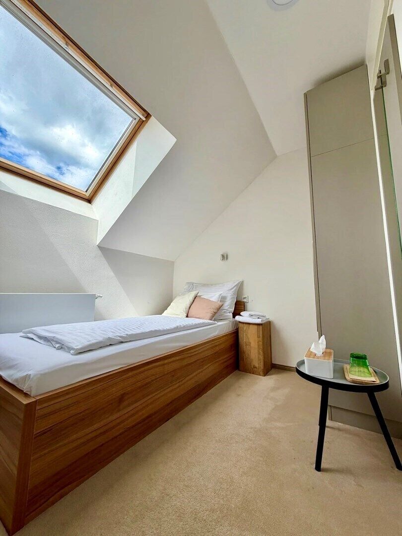 A bedroom with a sloped ceiling, a bed, a bedside table, and a small round table with a tissue box and green object.