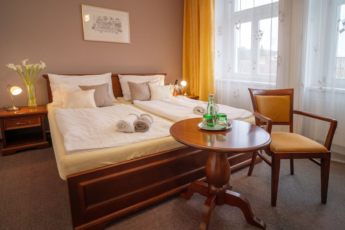 A hotel room with two beds, a round wooden table, a chair, a nightstand with a lamp, and a window with curtains.