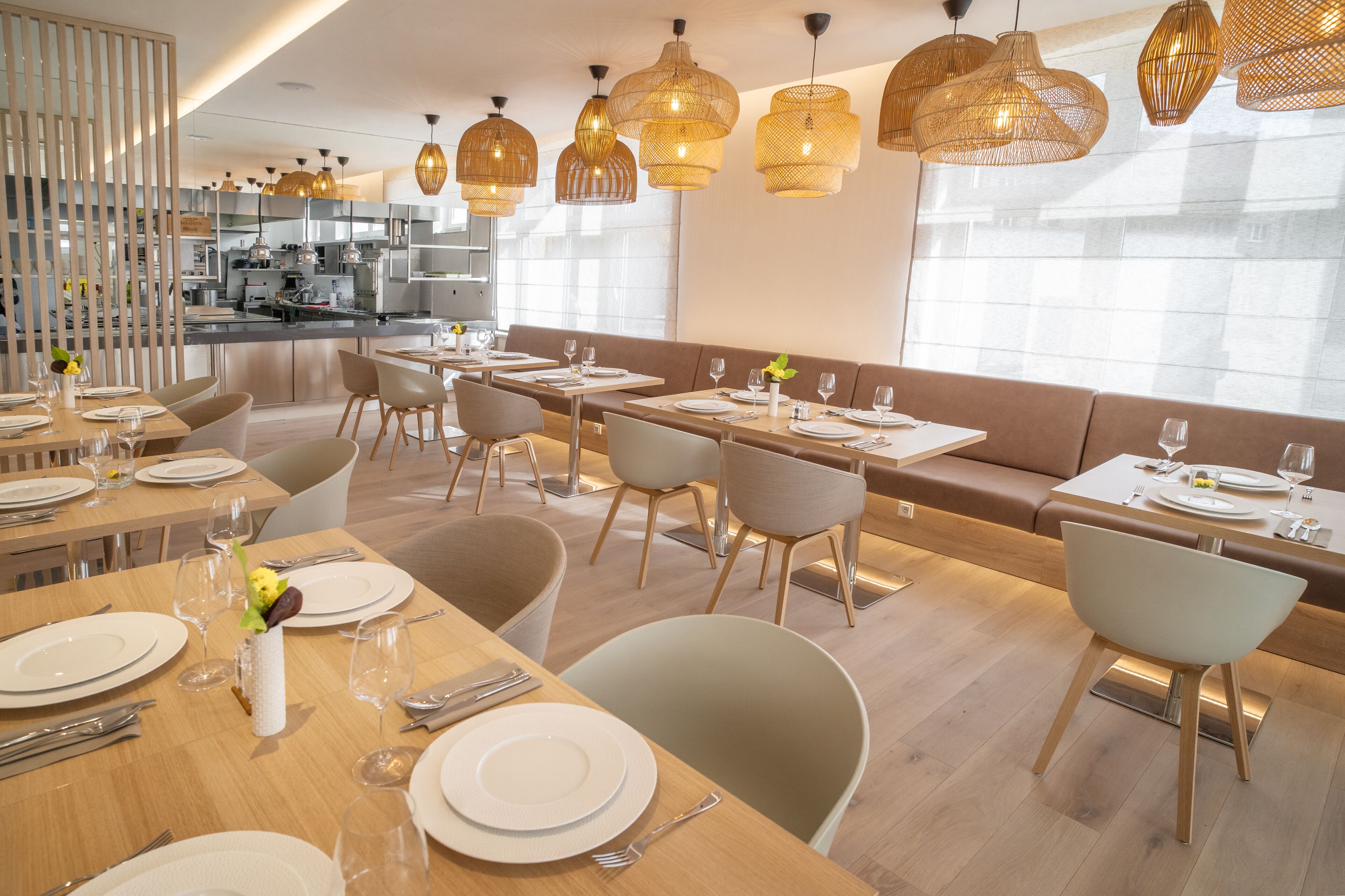 A modern dining area with multiple tables and chairs, a kitchen in the background, and large windows allowing natural light.