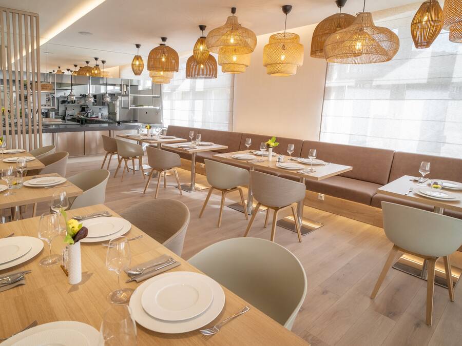 A modern dining area with multiple tables and chairs, a kitchen in the background, and large windows allowing natural light.