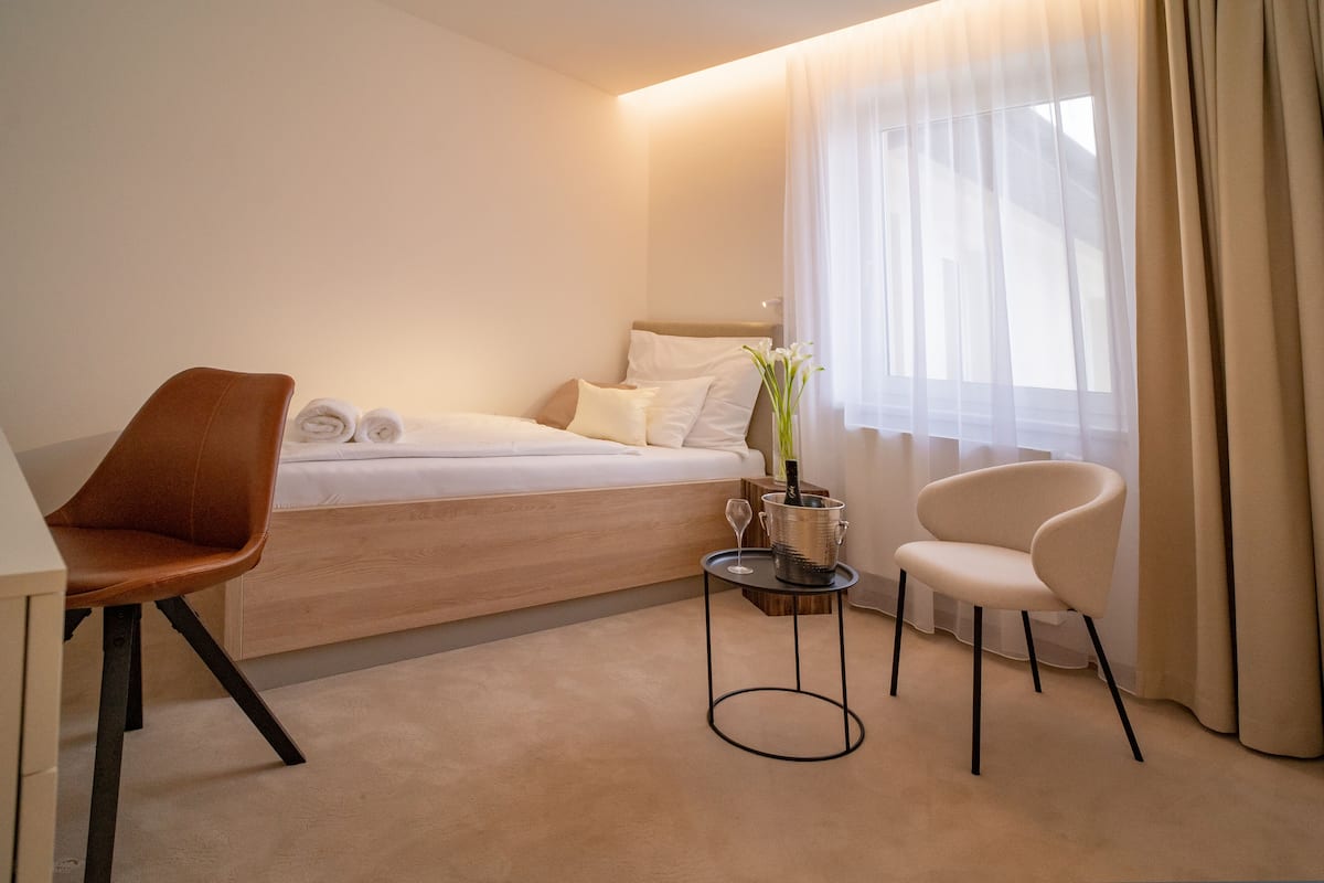 A hotel room with a bed, two chairs, a small table with a wine glass and a vase, and sheer curtains.