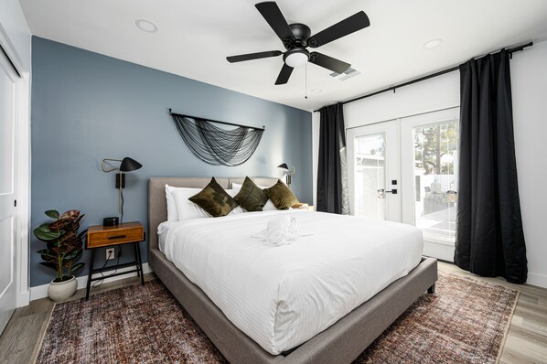 Een moderne slaapkamer met een bed, een plafondventilator, een nachtkastje met een lamp en uitzicht op de omgeving door een groot raam.