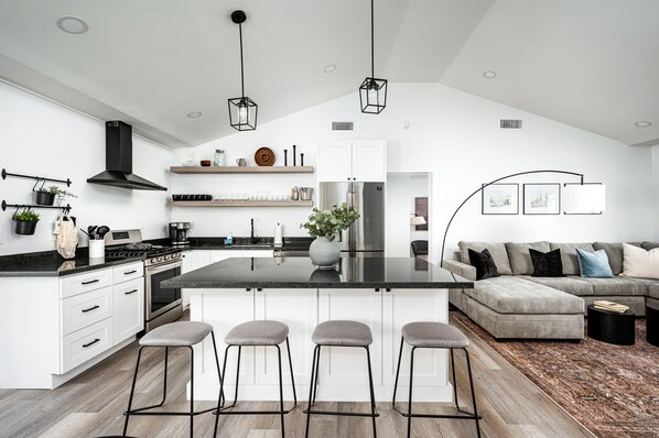 A modern kitchen with white cabinetry, a central island with bar stools, a grey sofa, and a black ceiling fan. - Swim up to cooking & entertainment, play corn hole, and relax by the fire at Cascada (Scottsdale)