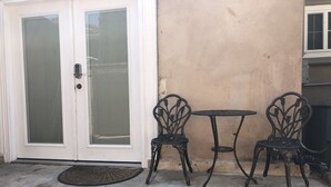 Une terrasse avec deux chaises en métal et une petite table, une porte blanche avec un panneau en verre et un mur de couleur beige.