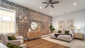 A modern living room with a brick wall, a ceiling fan, a sofa, a bed, and a side table with a lamp.
