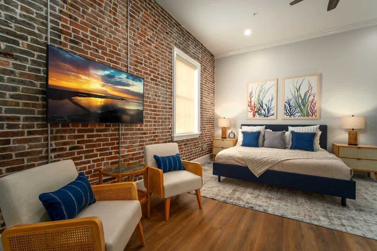 A bedroom with a brick wall, a bed, two nightstands, a TV, and a window.
