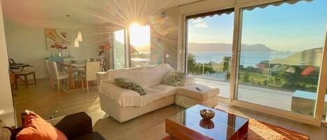 A modern living room with a white sofa, a glass coffee table, and a view of the sea.
