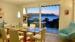 Een eetgedeelte met een glazen tafel en stoelen, een telescoop, en uitzicht op zee door grote ramen.