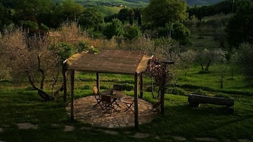 Een rustieke houten pergola met een stenen vloer en metalen stoelen, omgeven door weelderige begroeiing en een uitzicht op glooiende heuvels in de verte.