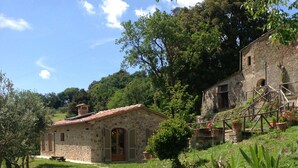 A stone building with a tiled roof, a stone pathway leading to the entrance, and lush greenery surrounding the property. - Tuscan country house with enchanting view and swimming pool (Radicondoli)