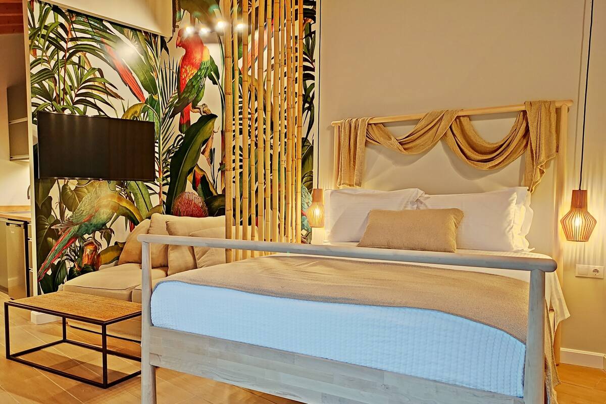 Un dormitorio con cama, mesita de noche, lámpara y una pared con un mural tropical.