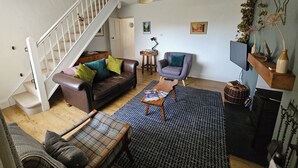 A living room with a brown leather sofa, a plaid armchair, a wooden coffee table, a blue patterned rug, a grey armchair, a small side table, a television, and a staircase with a white railing. - Cheerful two bedroom cottage in the Forest of Dean (Ruardean)