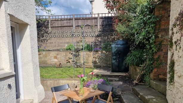 A small patio with a wooden table and chairs, a vase with flowers, and a garden view. - Cheerful two bedroom cottage in the Forest of Dean (Ruardean)