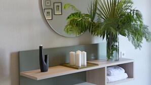 Une salle de bain moderne avec un miroir rond, une étagère murale agrémentée d’une plante, un vase noir et un tabouret à motifs.