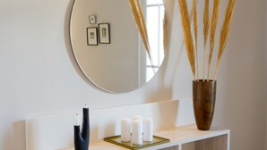 Une salle de bain moderne avec un miroir rond, un luminaire fixé au mur, un meuble-lavabo en bois avec un miroir, un vase contenant des plantes séchées et un tabouret à motifs.
