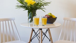 Une petite table ronde est agrémentée d’un vase de fleurs jaunes, de deux verres de jus d’orange et d’un saladier de fruits. Deux chaises blanches sont placées de part et d’autre de la table.