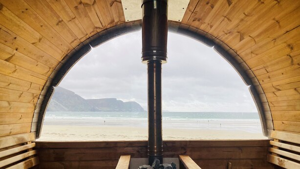 Eine Holzdiele mit einem großen Bogenfenster mit Blick auf einen Strand. Im Inneren befindet sich ein Kaminofen und eine kleine Sitzecke.
