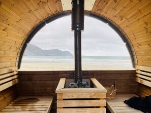 Una cabaña de madera con un amplio ventanal arqueado que da a una playa. En el interior, hay un estufa de leña y una pequeña zona de estar.
