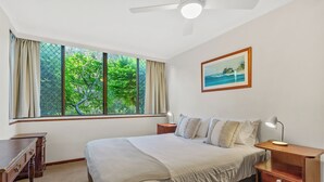Un dormitorio con cama, mesitas de noche, un escritorio, un ventilador de techo y una ventana con cortinas.